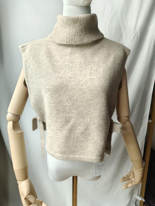 Gilet in maglia beige senza maniche con collo alto e lacci laterali, esposto su manichino. Capo in morbida lana a maglia fine, stile minimalista chic. Perfetto come maglione smanicato versatile per look casual e formali. Dettagli con fiocchi laterali