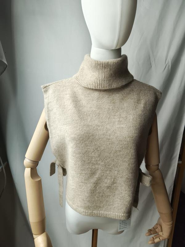 Gilet dolcevita senza maniche in maglia color beige avena con lacci laterali regolabili, esposto su manichino. Capo in tricot dal design minimalista e raffinato, ideale per layering autunnale. Perfetto come campione per acquisti all'ingrosso di magli
