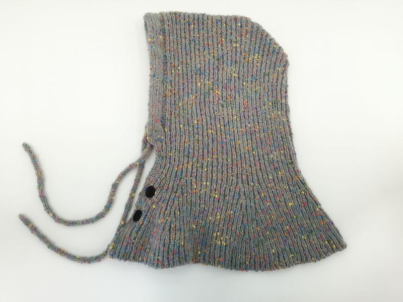 Cuffietta neonato in maglia spigata grigia multicolore con laccetti sottogola e due bottoni a pressione neri, vista dall'alto su sfondo neutro. Accessorio invernale lavorato a maglia per bebè, ideale per completare outfit neonato con dettagli pratici