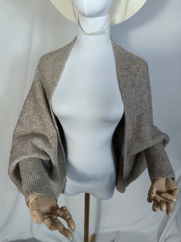 Shrug in maglia color taupe a maniche balloon con bordi a coste, modello aperto sul davanti esposto su manichino. Cardigan corto da donna in tricot elasticizzato, ideale come capospalla leggero per look casual o layering moda ingrosso.